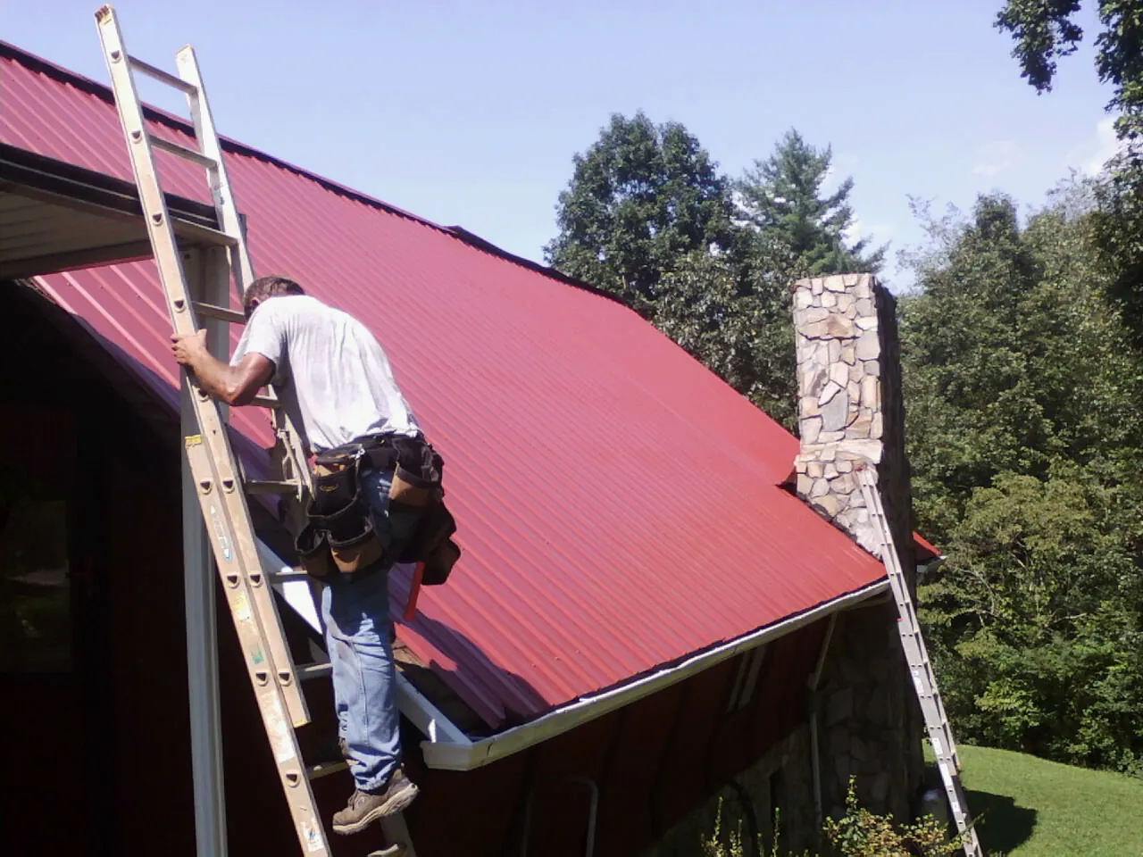 Roofer on a ladder beside a red metal roof and stone chimney — Soffit & Fascia Repair by Roofing Brothers in Belvidere